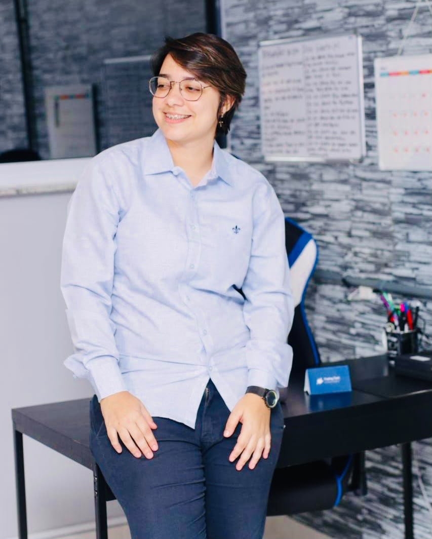 Gessica de camisa azul, sentada na borda da mesa do escritório.